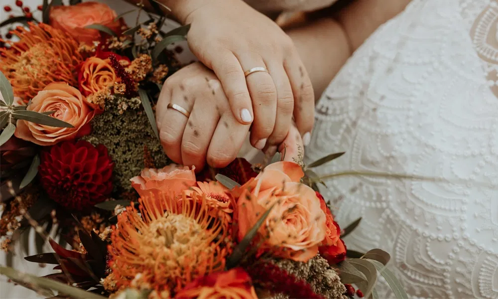 Zwei Hände mit Trauringen liegen auf einem farbenfrohen Brautstrauß aus Orange- und Rottönen.