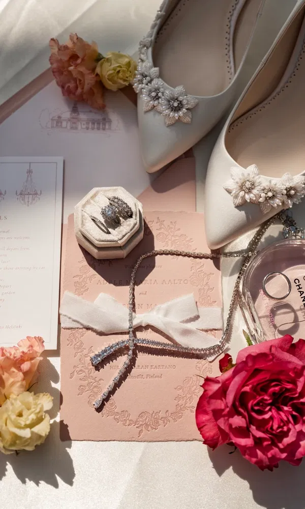 Elegantes Flatlay mit Brautschuhen, Diamantringen, Einladung und Chanel-Parfum, dekoriert mit Rosen.