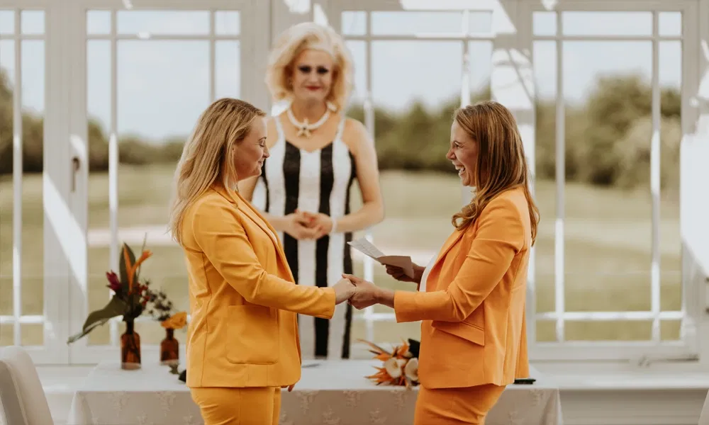 Zwei Frauen im Partnerlook mit orangefarbenen Anzügen stehen bei ihrer Trauung Hand in Hand, sichtbar glücklich und gerührt.