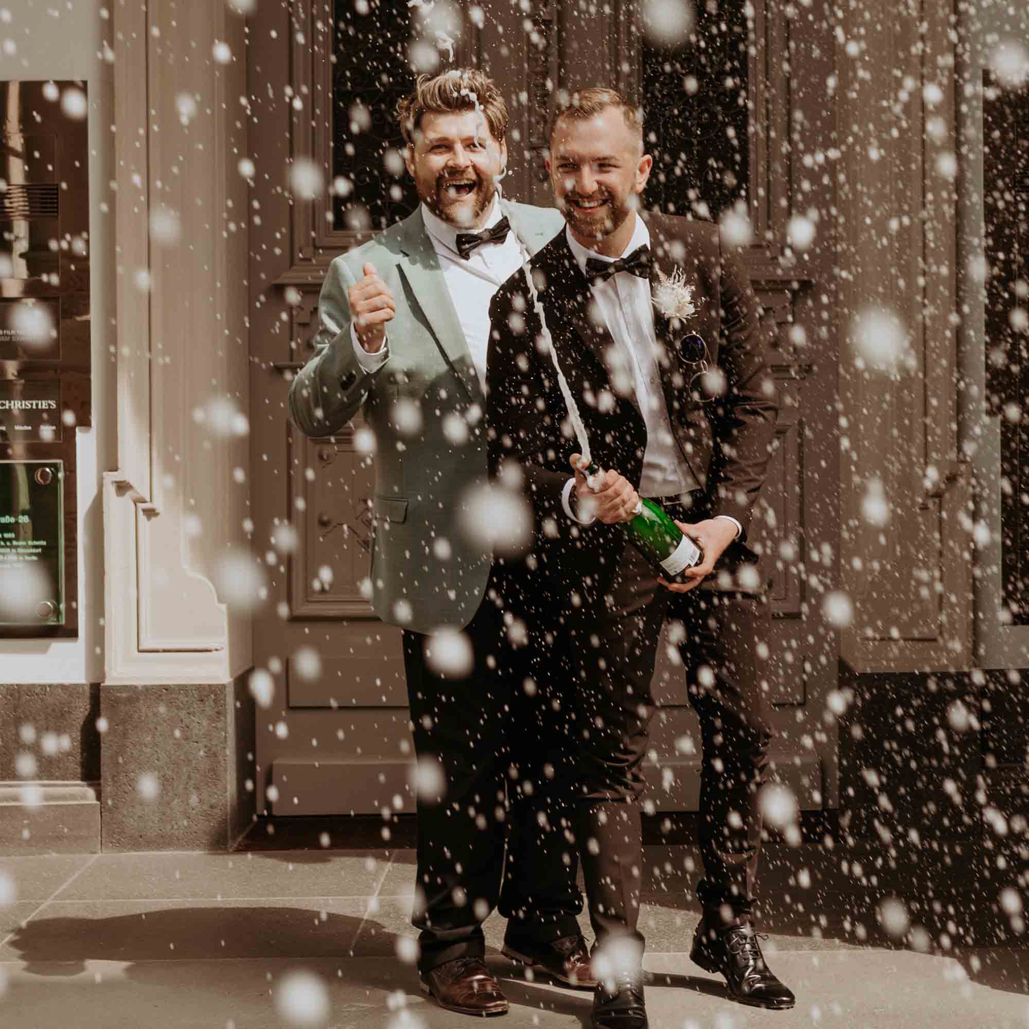 Frisch verheiratetes Paar in Anzügen lacht und sprüht Champagner, während perlender Schaum die Szene umhüllt.