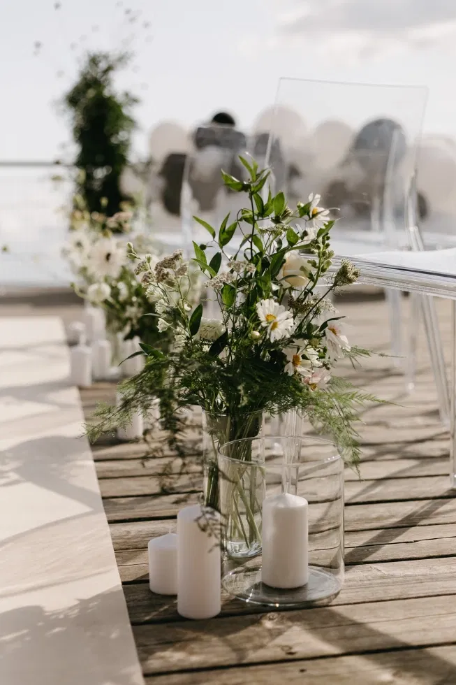 Zarte Blumenarrangements in Glasvasen mit weißen Kerzen entlang eines Holzstegs bei einer freien Trauung