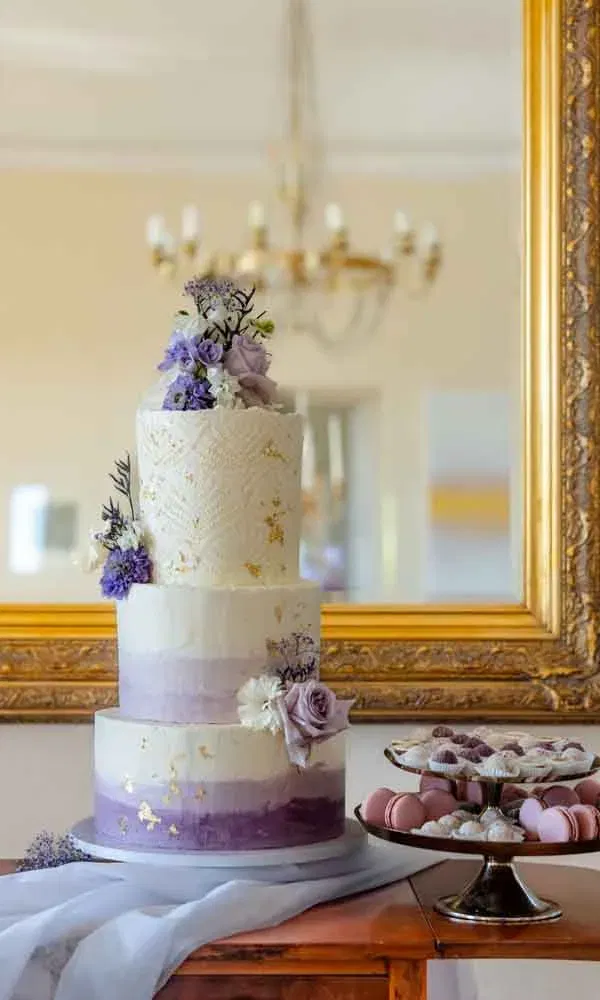 Dreistöckige Hochzeitstorte in Weiß-Lila mit Goldakzenten und Blumendekoration, daneben Macarons und Pralinen