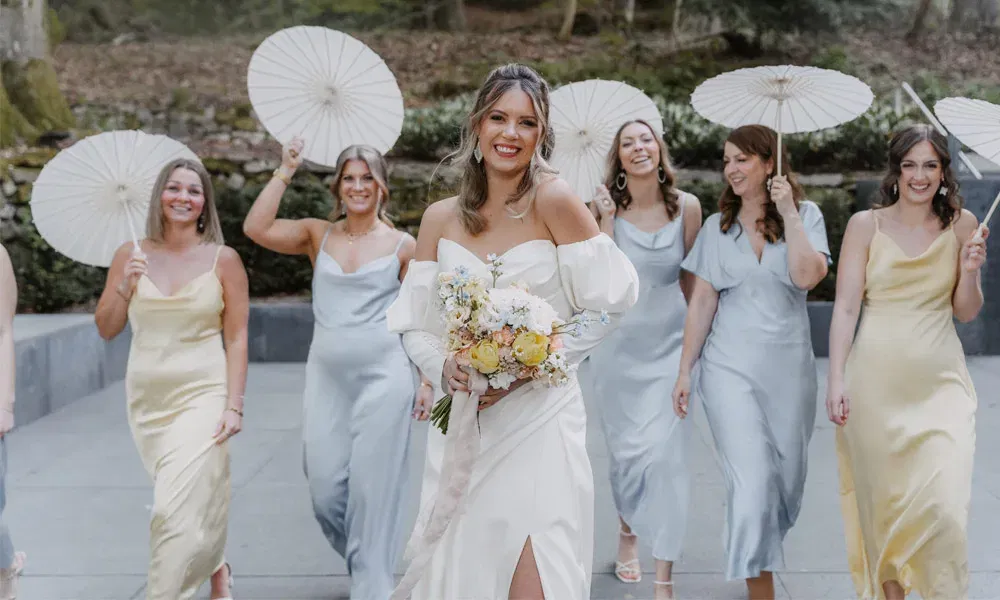 ChatGPT: Braut in elegantem weißen Kleid mit Blumenstrauß, umgeben von Brautjungfern in hellblauen und gelben Kleidern, die weiße Sonnenschirme tragen.