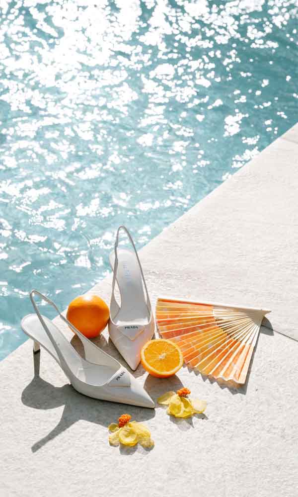 Weiße Prada-Slingback-Heels am Pool mit Orange, Fächer und Blüten als sommerliche Hochzeitsinspiration.