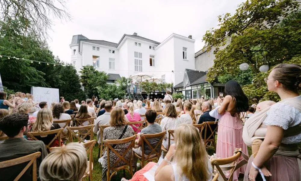 Gäste sitzen im Garten der „Villa Lindström“ am Niederrhein und verfolgen die feierliche Eröffnung mit Modenschau vor der historischen Villa.