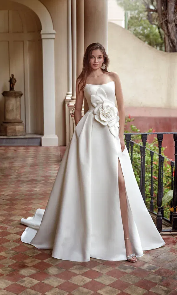 Zeitloses Brautkleid mit langer Schleppe, auffälliger Blume und femininem Schlitz für einen glamourösen Look.