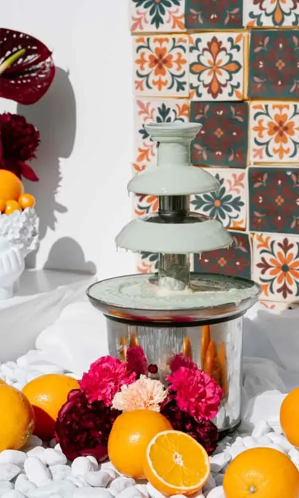 Eleganter Schokoladenbrunnen, dekoriert mit frischen Orangen und bunten Blumen vor mediterranen Fliesen.