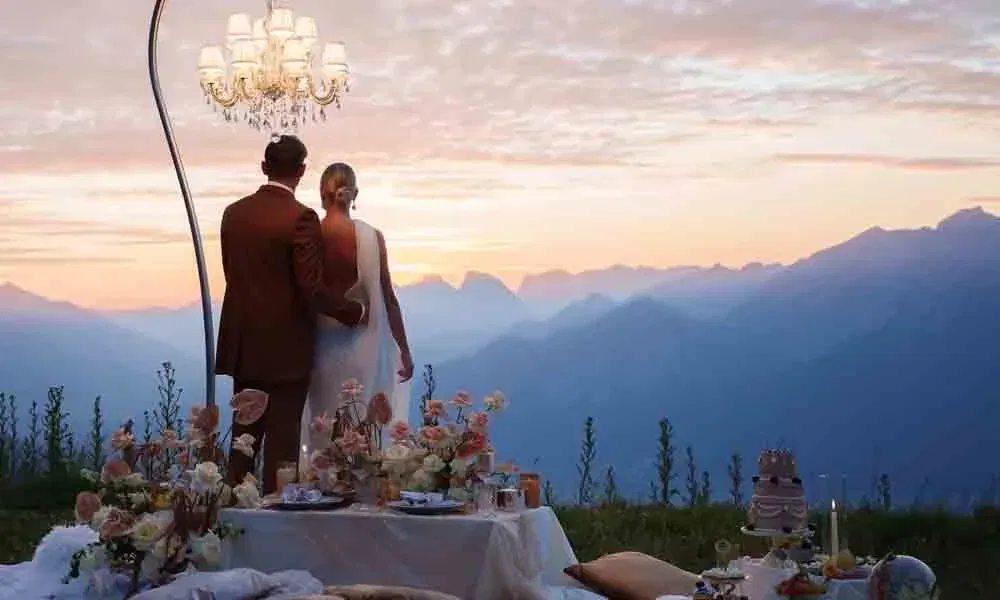Hochzeitspaar steht Arm in Arm vor alpiner Bergkulisse, festliche Picknicktafel mit Blumen und Kerzen im Vordergrund.
