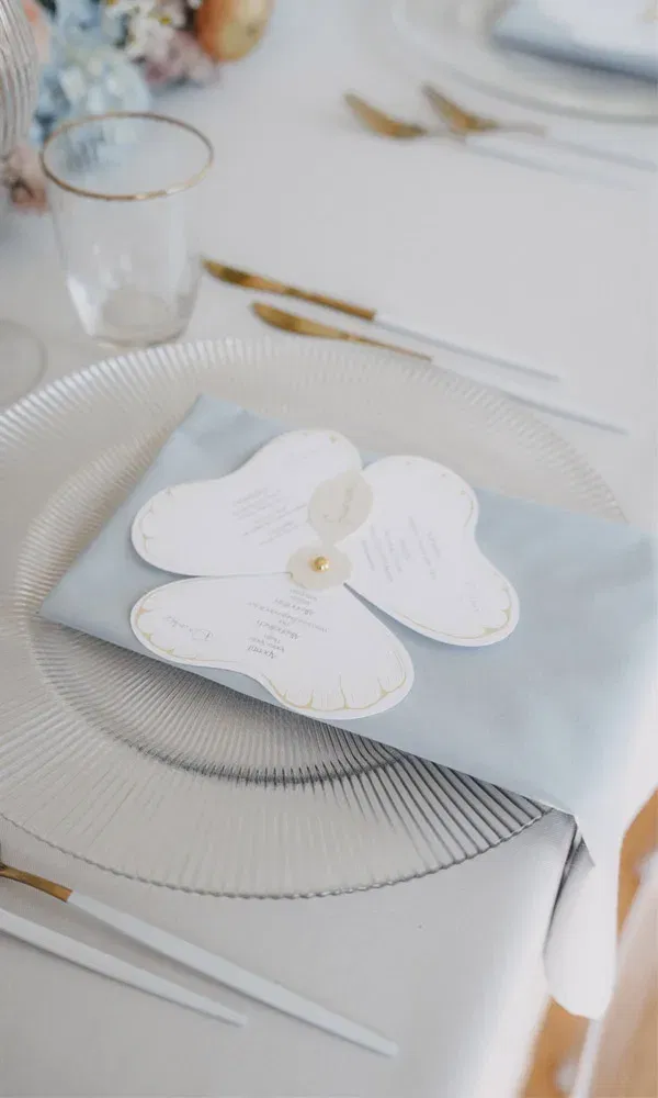 Elegante Hochzeitstafel mit zartblauer Serviette, darauf eine blütenförmige Menükarte mit goldenen Akzenten.