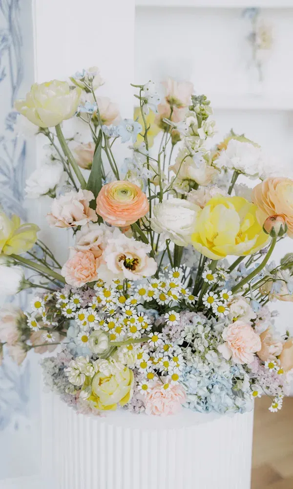 Farbprächtige Blumendekoration in Pastelltönen mit Ranunkeln, Margeriten, Hortensien und gelben Tulpen in einer weißen Vase.