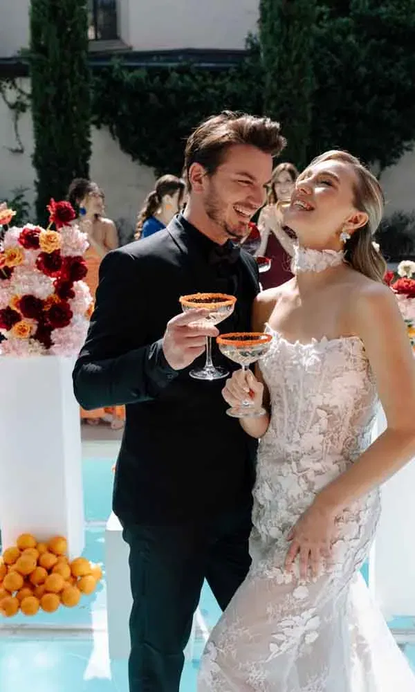 Braut und Bräutigam stoßen bei einer sommerlichen Hochzeit mit bunten Signature-Drinks fröhlich an.