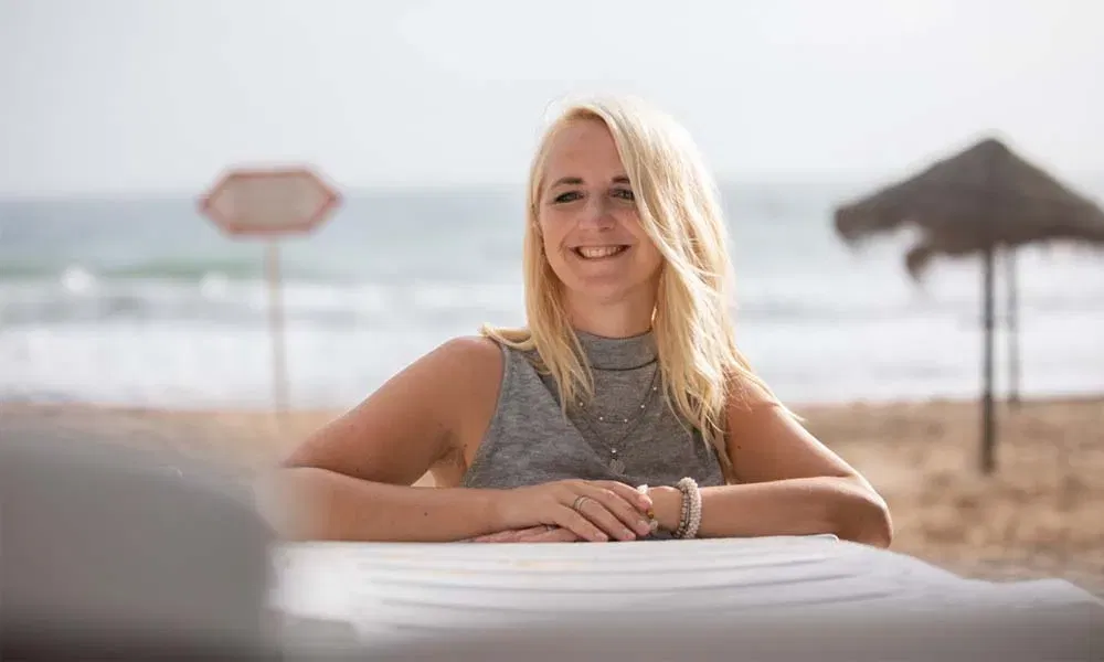 Entspannte Strandaufnahme: Frau sitzt an weißem Tisch, blickt freundlich zur Kamera; Küste und Schirm-Silhouetten verschwimmen.