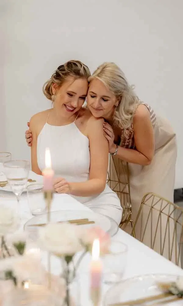 Emotionaler Augenblick zwischen Braut und Begleiterin, minimalistische Deko mit Glasgeschirr und Blumen.