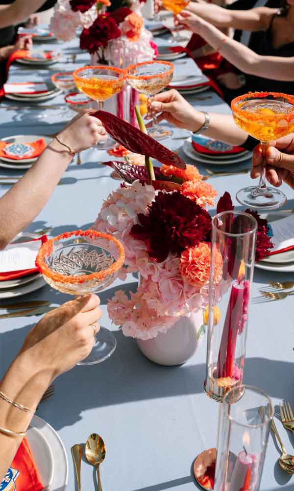 Hochzeitsgäste heben Gläser mit farbenfrohen Cocktails an einer festlich gedeckten Sommertafel mit Blumenarrangements.