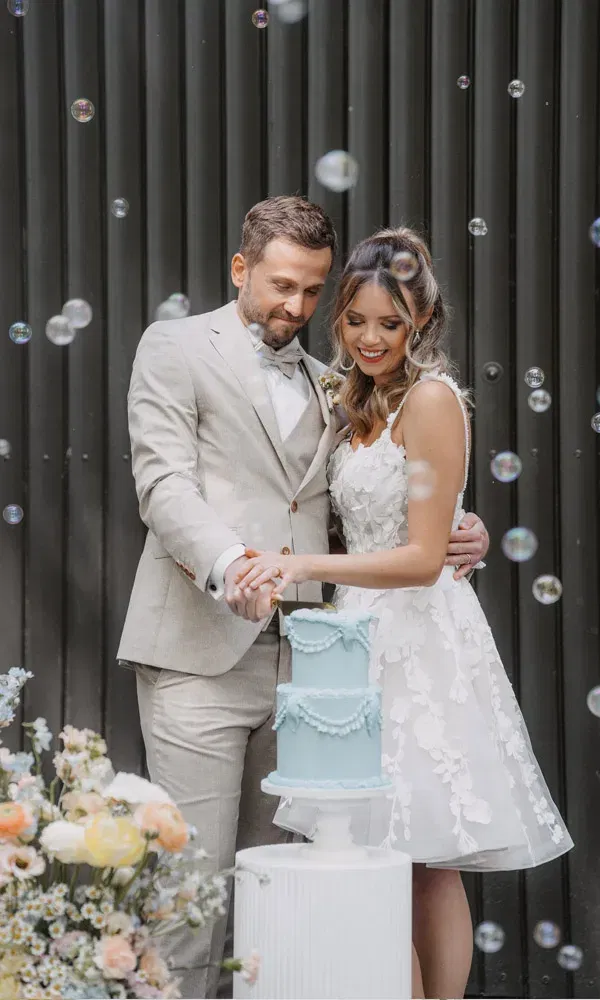 Brautpaar schneidet gemeinsam eine hellblaue Hochzeitstorte an, umgeben von Blüten und Seifenblasen.