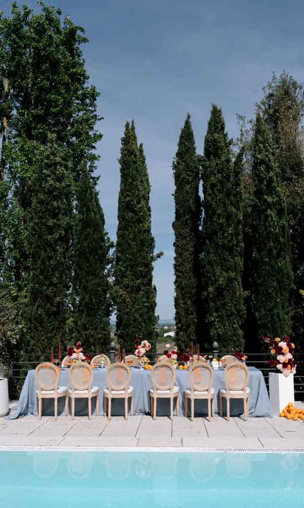 Sommerliche Hochzeitstafel mit blauer Tischdecke und floraler Dekoration vor hohen Zypressen am Pool.