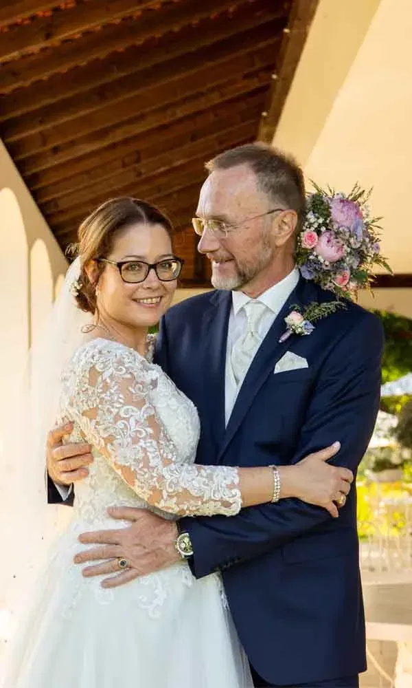 Brautpaar umarmt sich lächelnd, die Braut trägt ein Spitzenkleid mit langen Ärmeln und der Bräutigam einen dunklen Anzug mit Boutonniere.