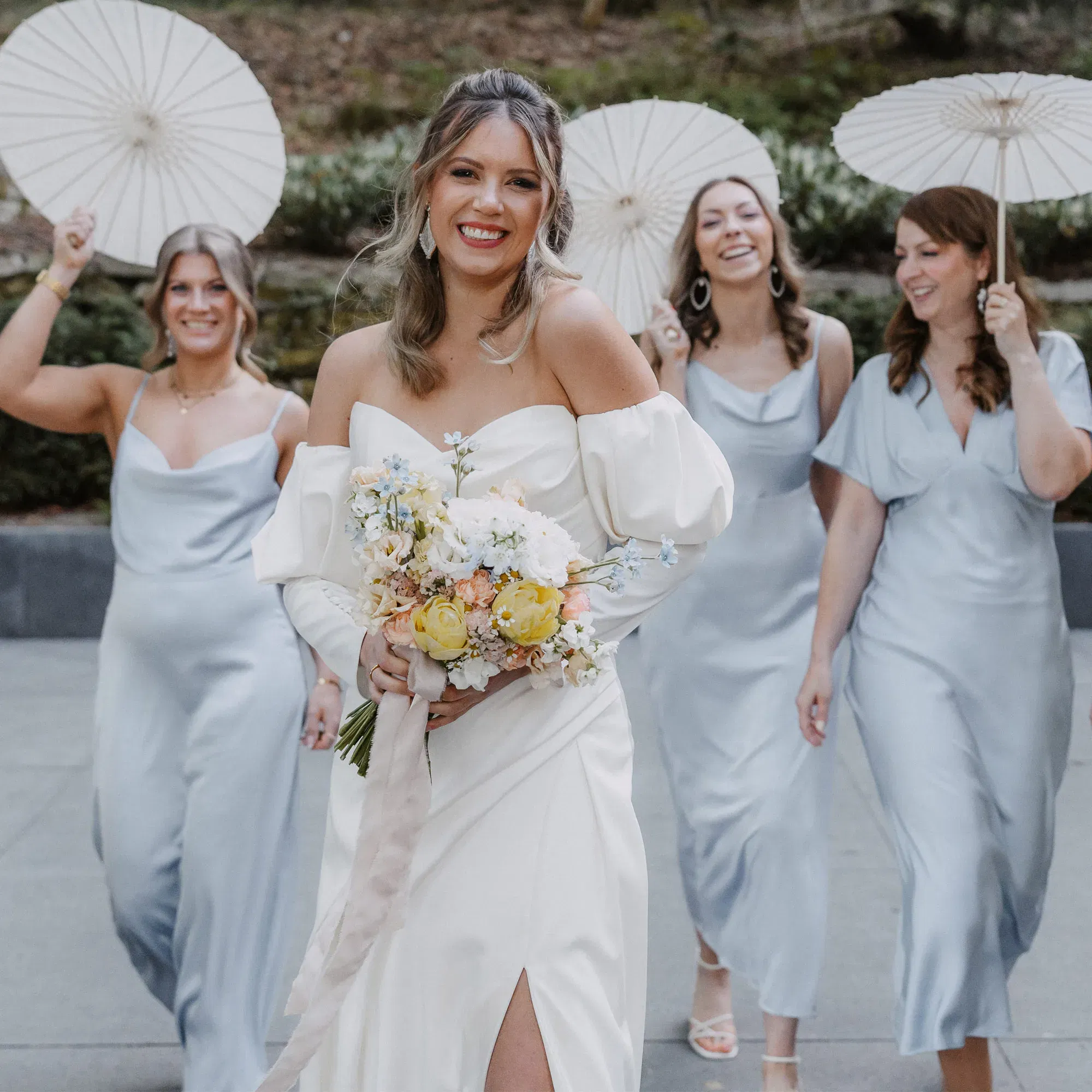 Braut in weißem Kleid mit Blumenstrauß, begleitet von drei Brautjungfern in hellblauen Kleidern mit weißen Sonnenschirmen.