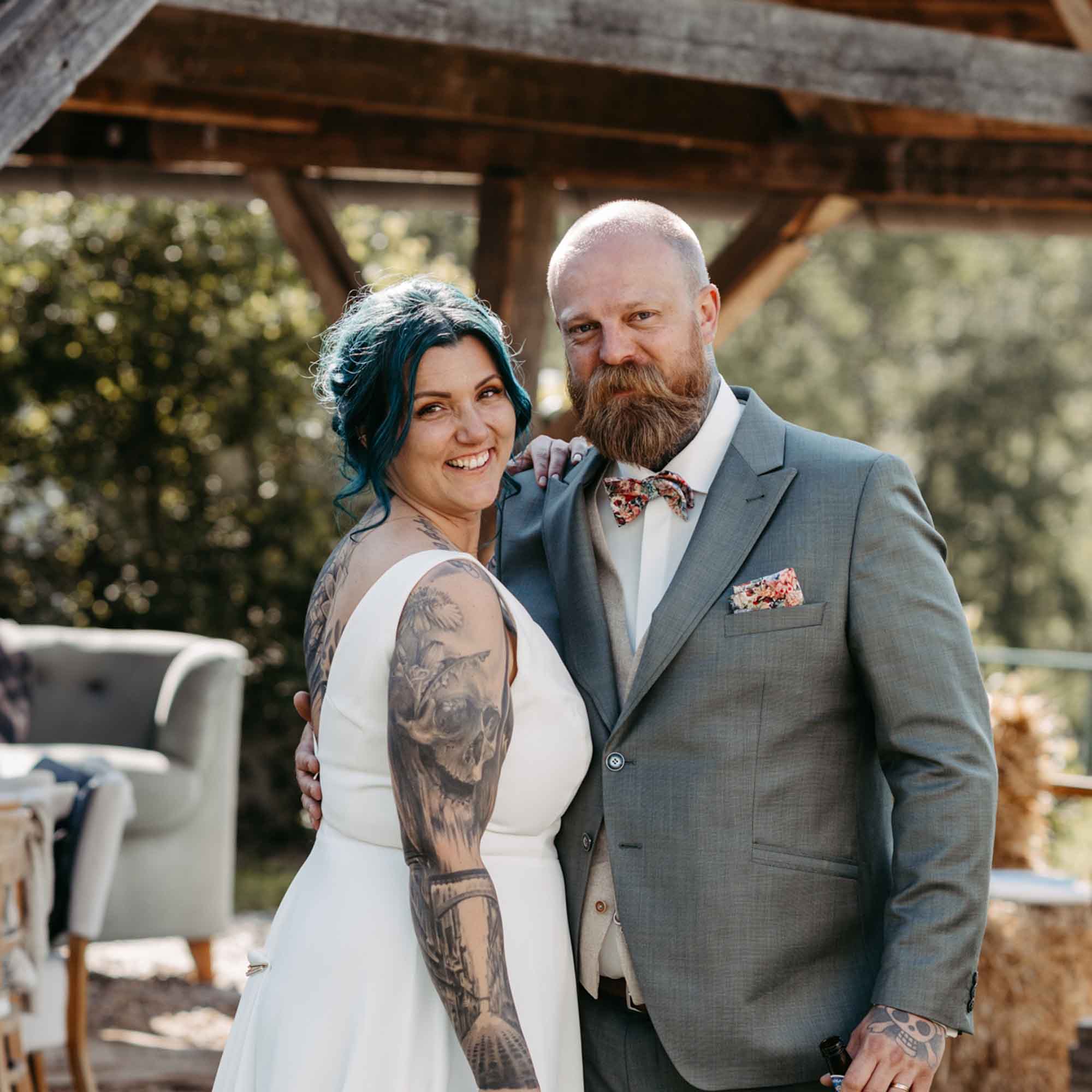 Braut mit tätowiertem Arm und blauem Haar steht lächelnd neben ihrem Bräutigam im grauen Anzug, beide posieren eng umschlungen im Freien.