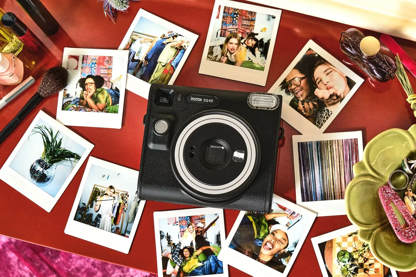 Fujifilm Instax SQ40 Sofortbildkamera liegt auf einem roten Tisch, umgeben von ausgedruckten Sofortbildern, Make-up-Utensilien und Accessoires.