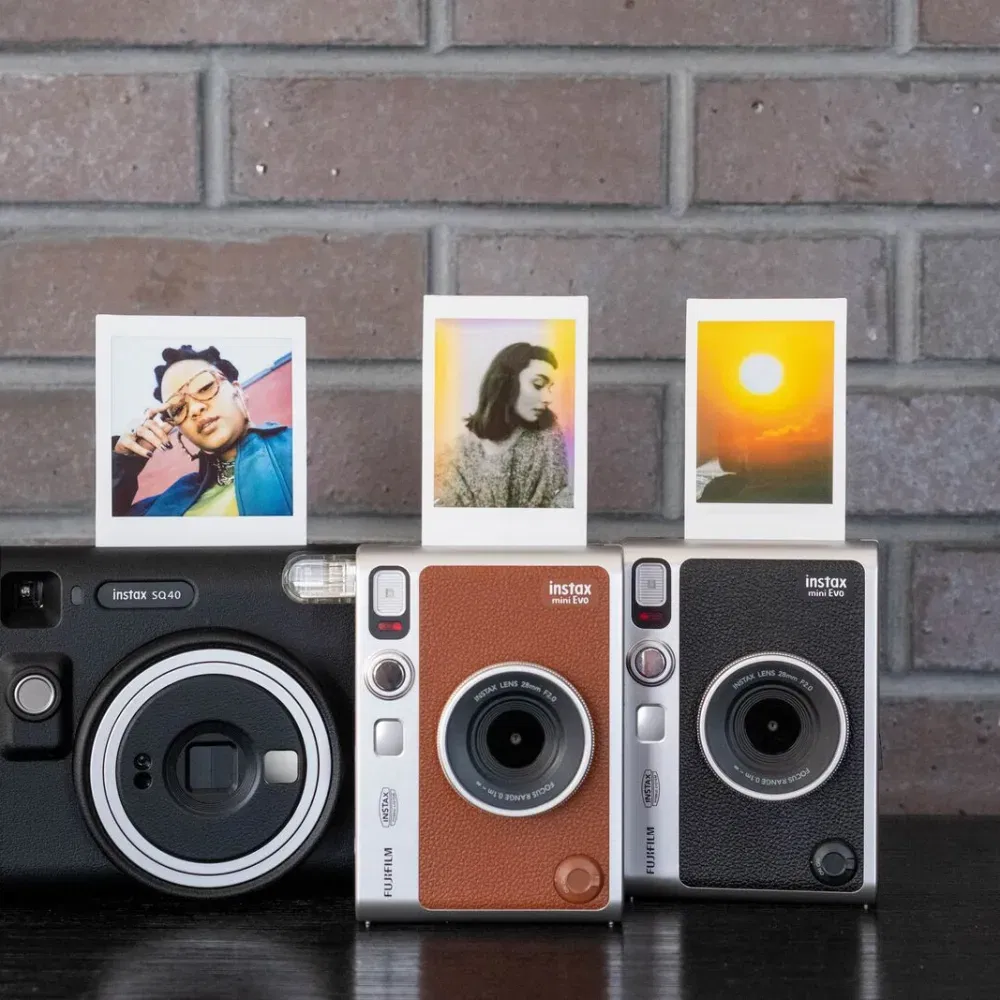Drei Fujifilm Instax Kameras – die schwarze SQ40 sowie zwei Instax mini Evo Modelle in Braun und Schwarz – stehen nebeneinander vor einer Ziegelwand, jede mit einem Sofortbild im Ausgabeschlitz.