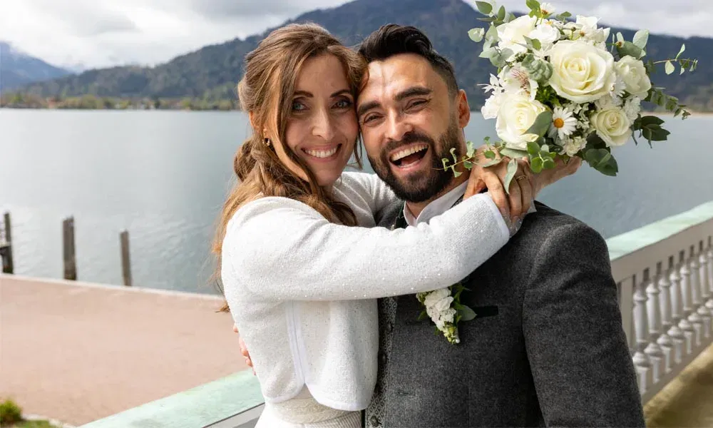 Strahlendes Brautpaar umarmt sich lachend vor malerischer See-Kulisse, die Braut hält einen Blumenstrauß mit weißen Rosen in die Höhe.