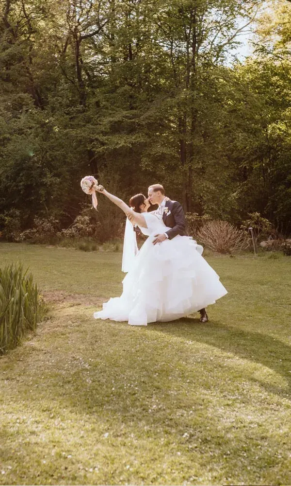 Bräutigam hebt seine Braut im Garten hoch und küsst sie, während sie den Brautstrauß jubelnd in die Luft hält.