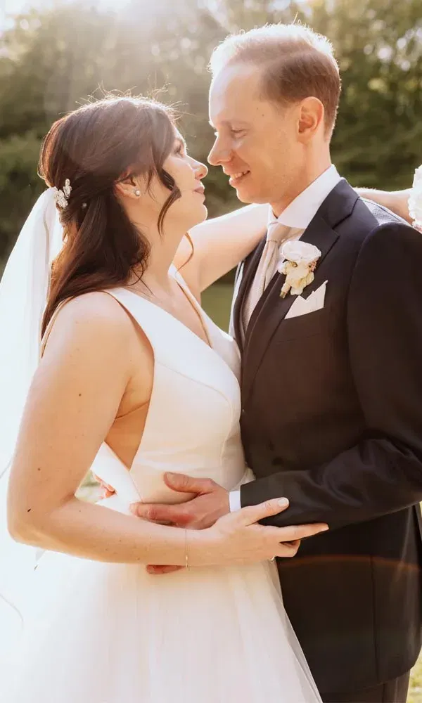 Brautpaar steht eng umschlungen im Sonnenlicht, schaut sich verliebt in die Augen und lächelt einander zu.