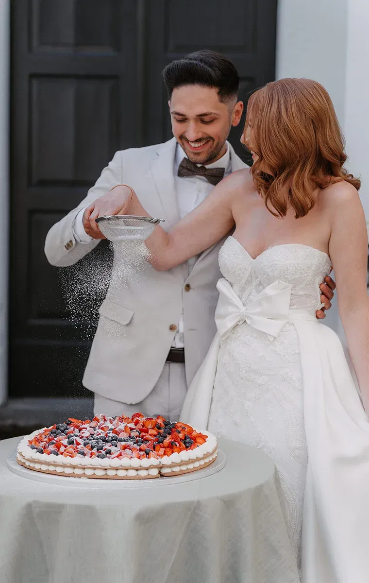 Braut und Bräutigam bestäuben ihre Hochzeitstorte lachend mit Puderzucker.