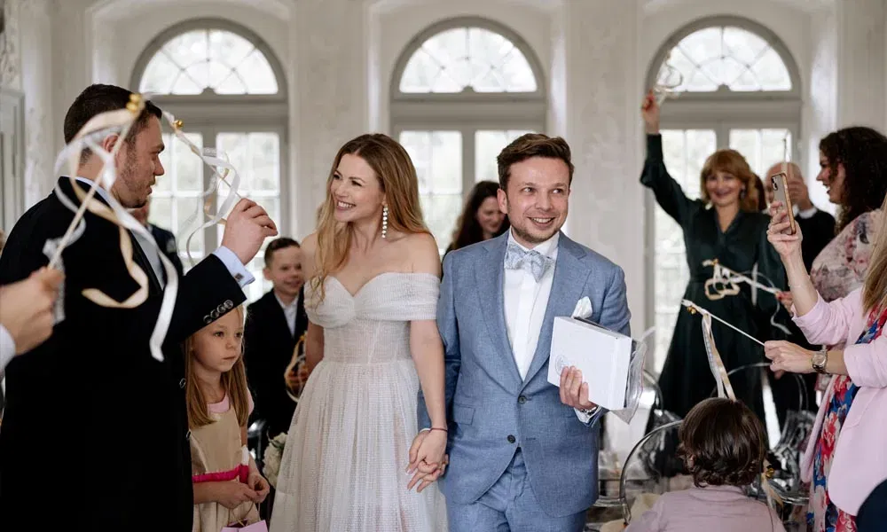 Frisch vermähltes Paar läuft lächelnd durch eine Hochzeitsgesellschaft, während Gäste mit Hochzeitswedeln jubeln und den Moment mit dem Handy festhalten.