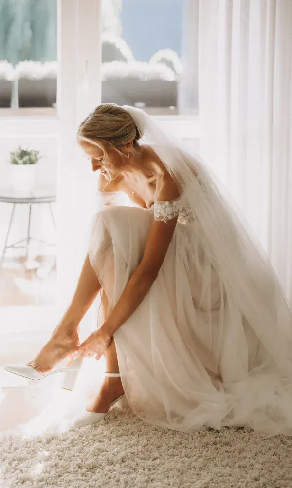 Zarte Momentaufnahme einer Braut, die sich im sanften Morgenlicht ihre Schuhe anzieht – ihr Tüllkleid und Schleier leuchten im Sonnenlicht.