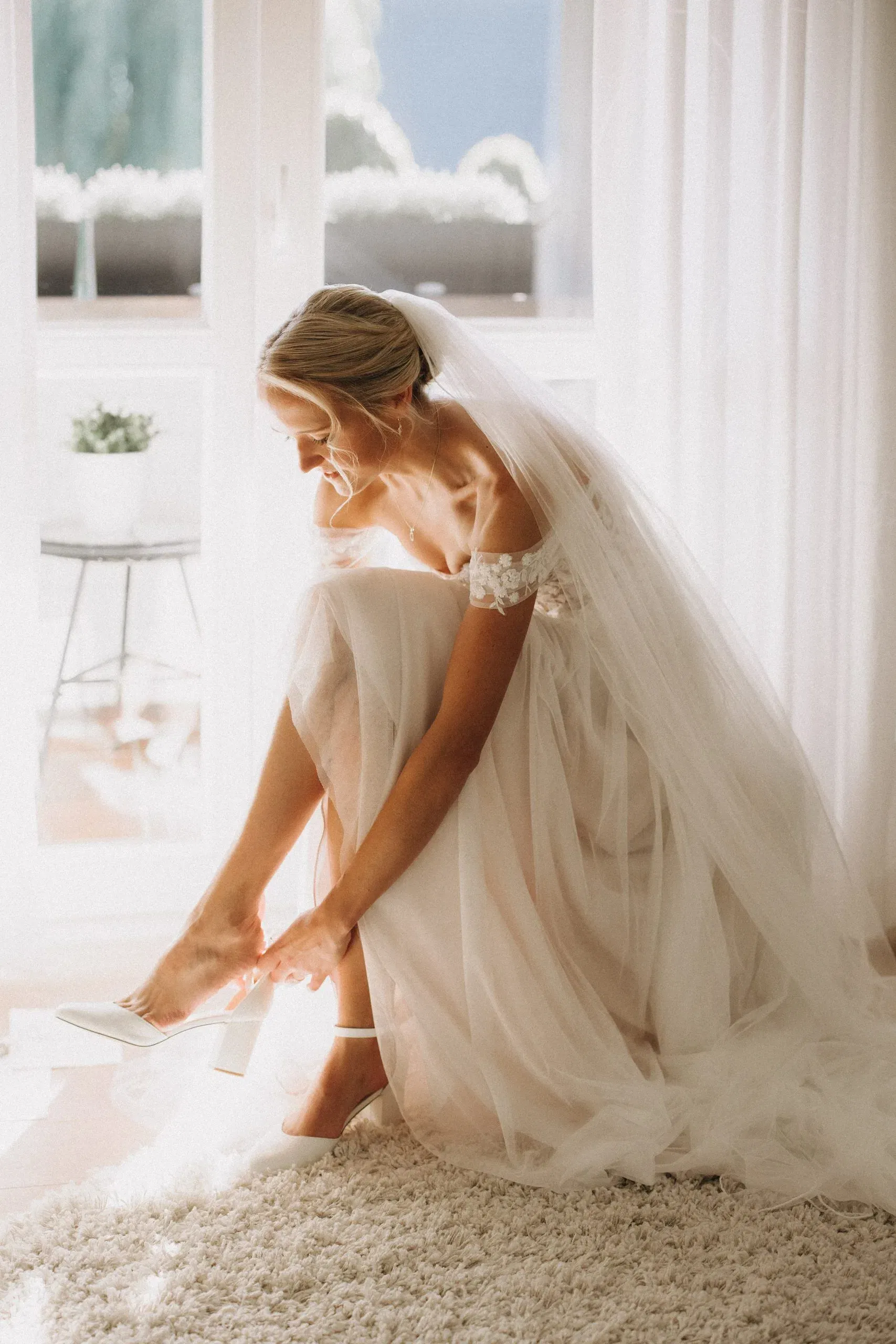 Zarte Momentaufnahme einer Braut, die sich im sanften Morgenlicht ihre Schuhe anzieht – ihr Tüllkleid und Schleier leuchten im Sonnenlicht.