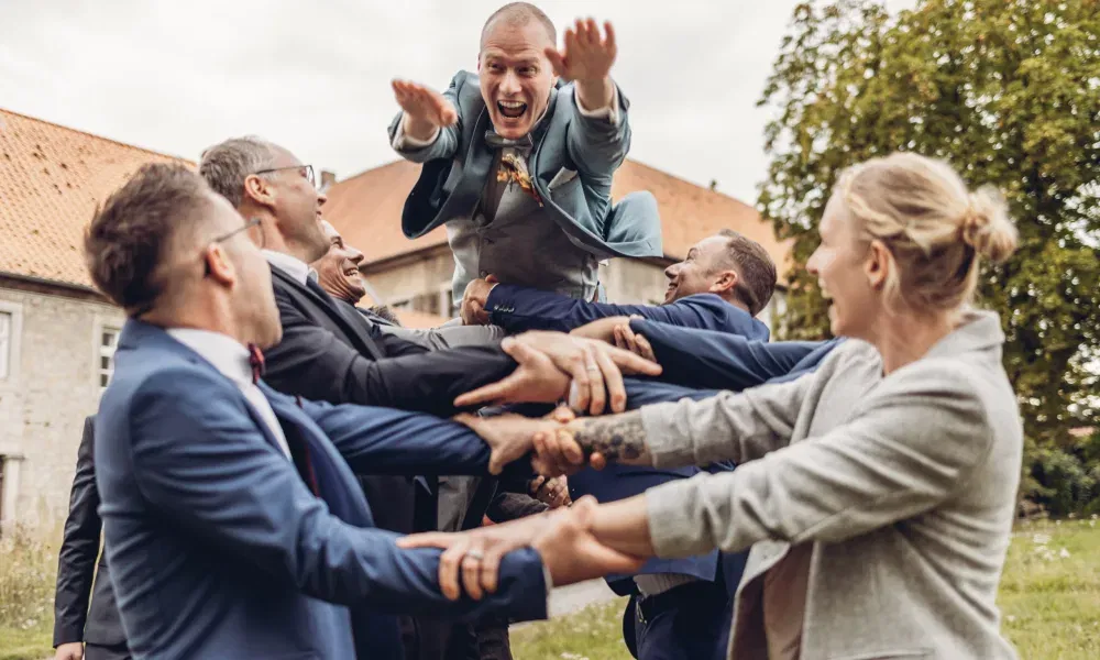 Fröhliche Hochzeitsgesellschaft wirft den Bräutigam lachend in die Luft – ein dynamischer Moment voller Freude und Gemeinschaft.