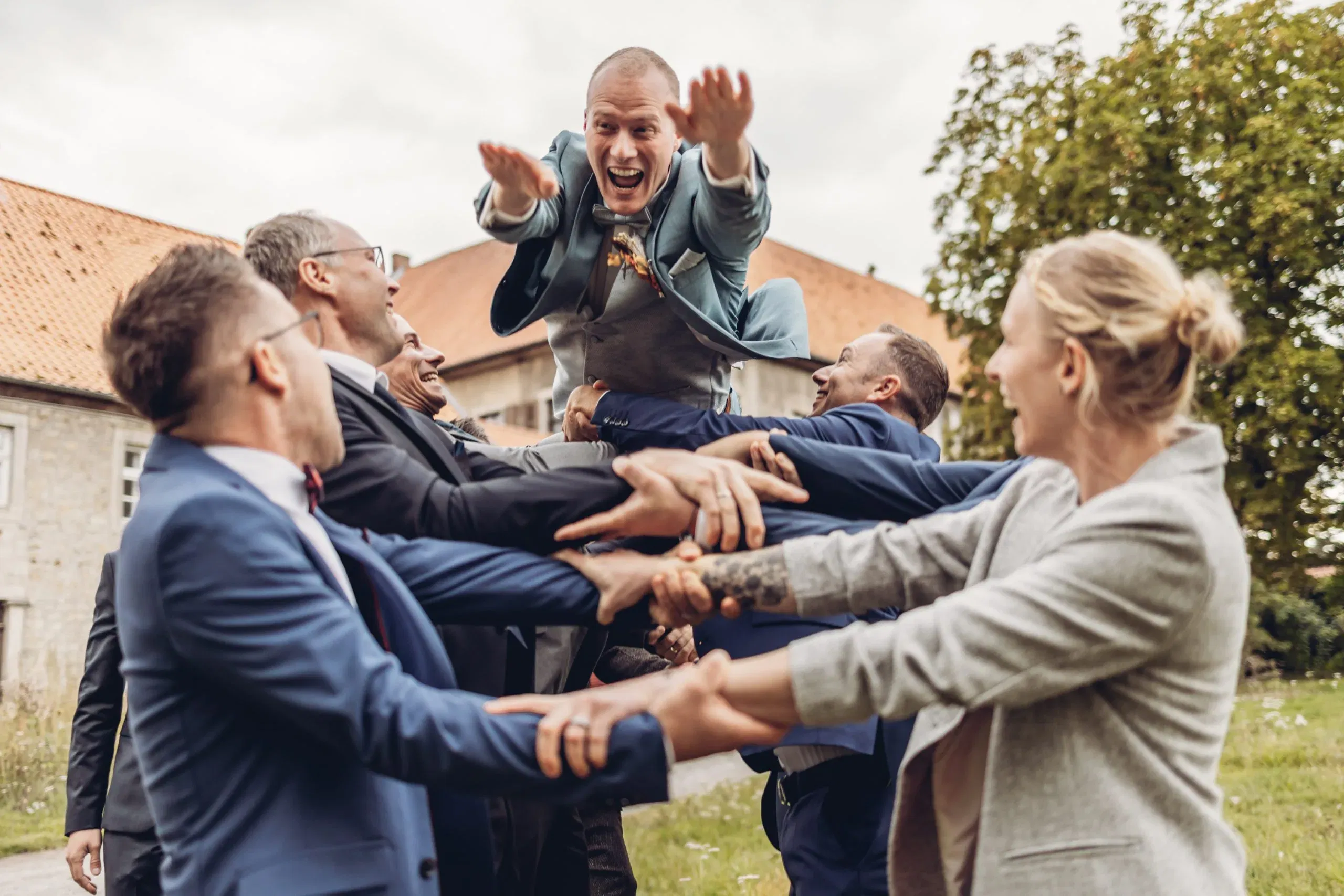 Fröhliche Hochzeitsgesellschaft wirft den Bräutigam lachend in die Luft – ein dynamischer Moment voller Freude und Gemeinschaft.