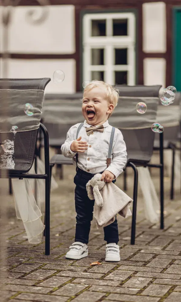 Lachender kleiner Junge in festlicher Kleidung spielt begeistert mit Seifenblasen – ein fröhlicher Moment während der Hochzeitsfeier.