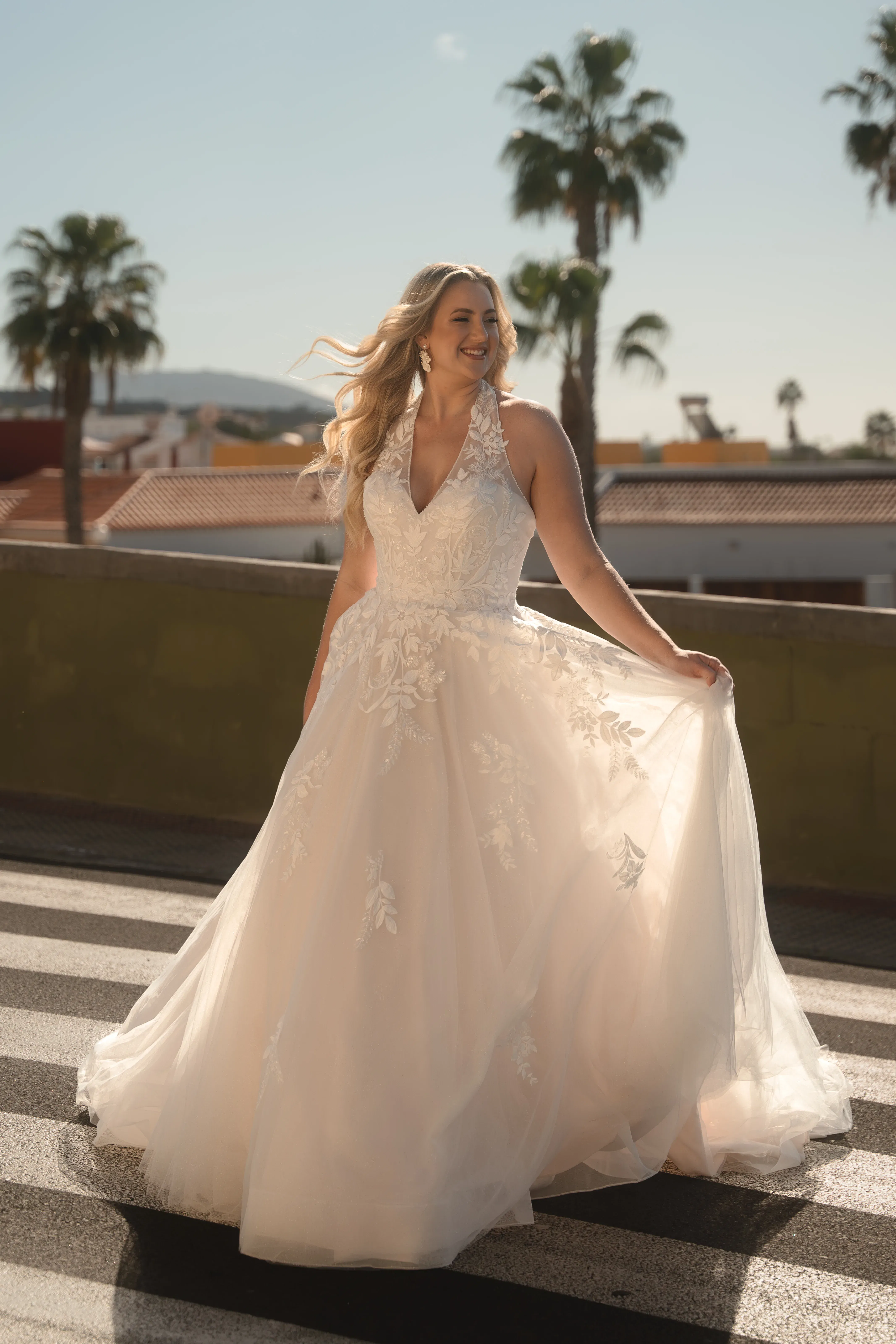 Plus-Size-Braut trägt ein fließendes A-Linien-Brautkleid mit floraler Spitze und tiefem V-Ausschnitt von Diane Legrand, lächelnd in der Sonne.