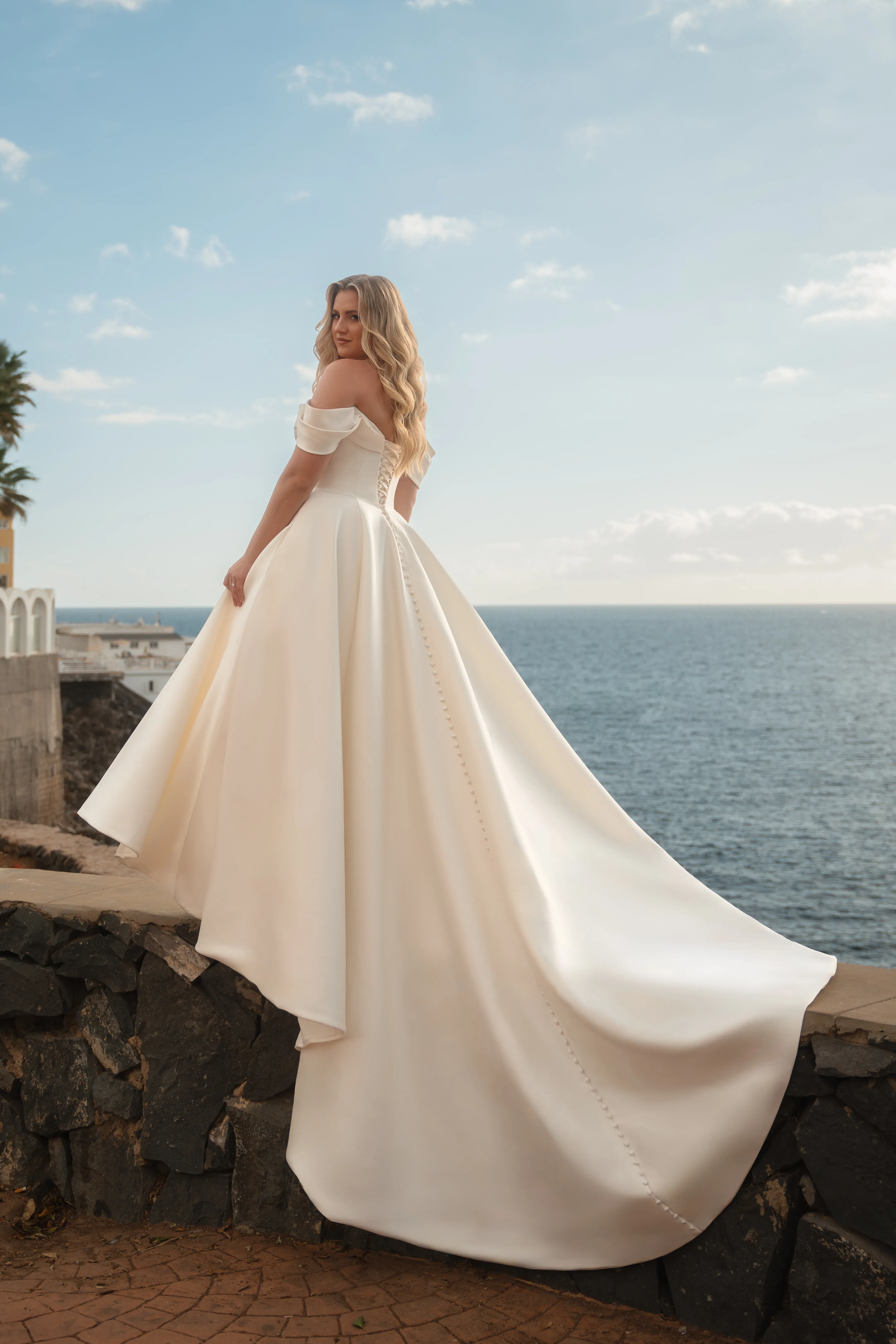 Elegantes Hochzeitskleid im Clean Chic mit abnehmbaren Off-Shoulder-Trägern und langer Schleppe, präsentiert von einer Plus-Size-Braut am Meer.