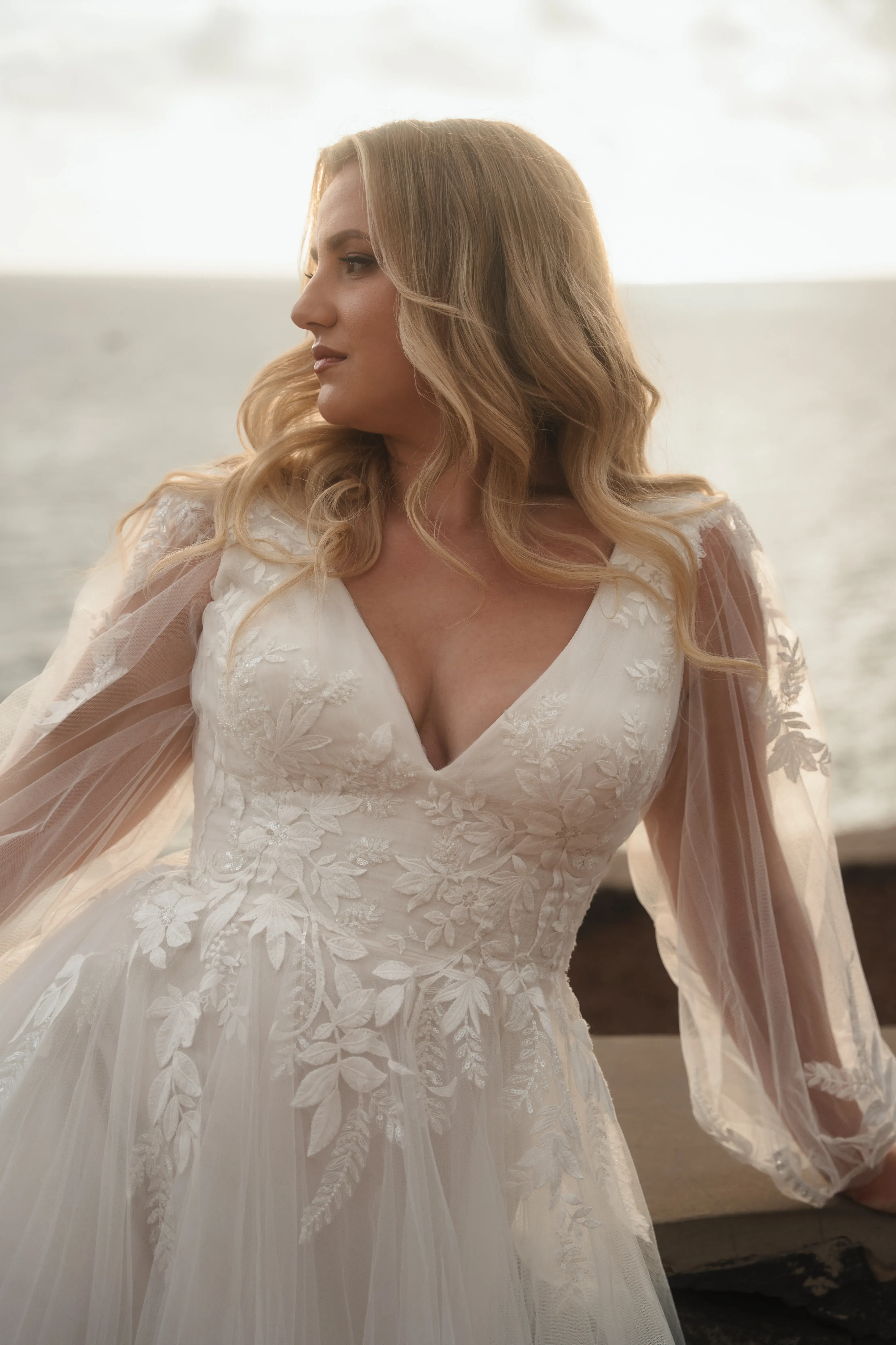 Zartes Curvy Brautkleid mit langen Tüllärmeln, V-Ausschnitt und floraler Spitze, perfekt für romantische Hochzeiten am Wasser.