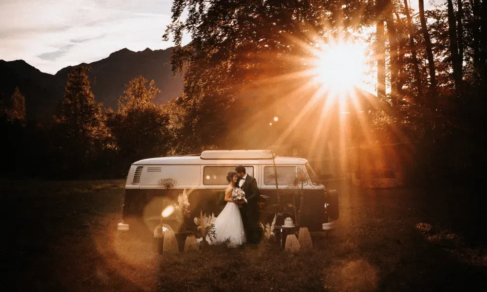 Romantisches Brautpaar steht innig vor einem Vintage-Bulli inmitten der Natur – goldenes Sonnenlicht taucht die Szene in warmen Abendglanz.