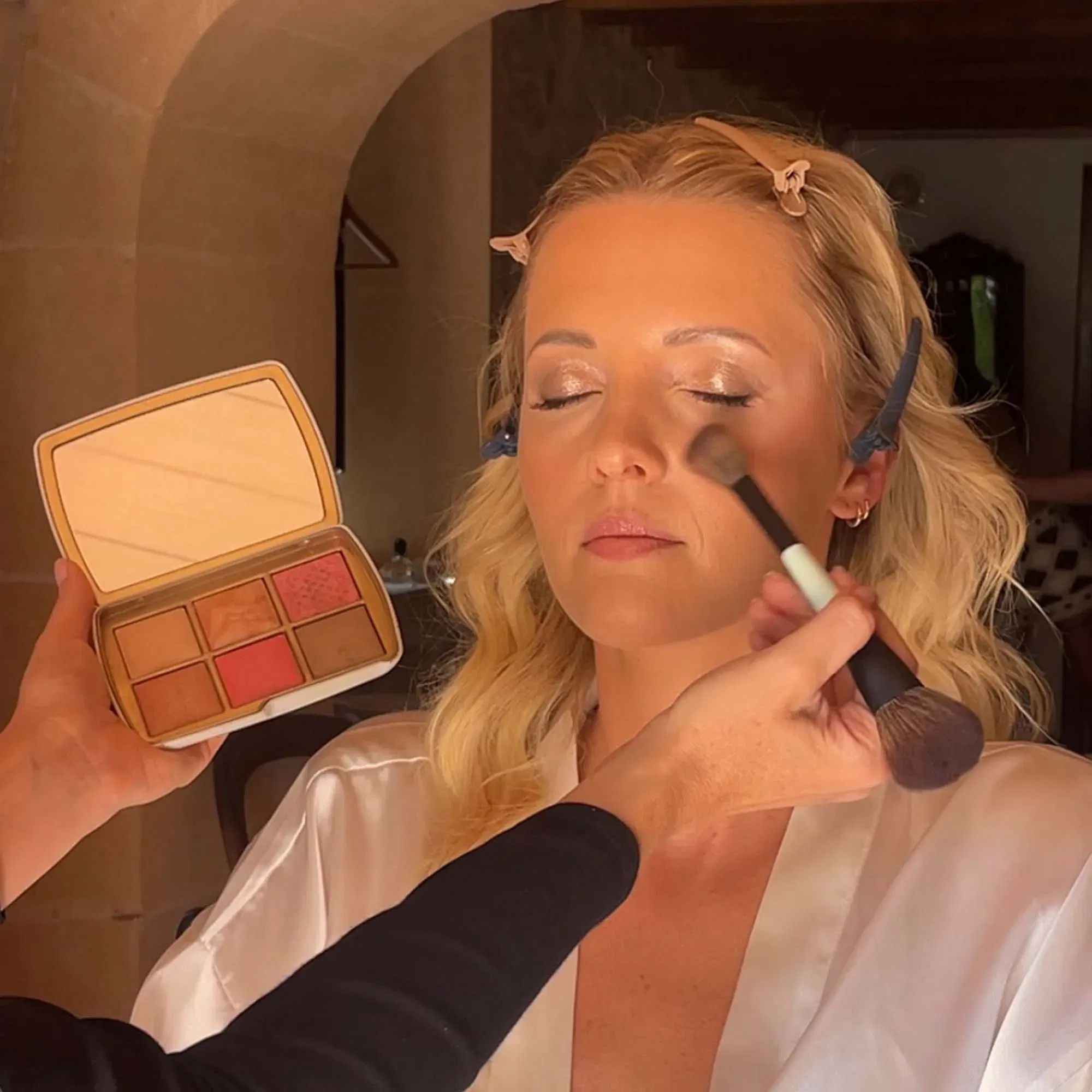 Visagistin trägt zart schimmerndes Make-up auf die Wangen einer Frau auf, die in einem Seidenmorgenmantel sitzt.