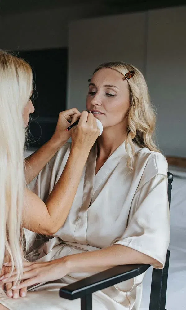 Frau sitzt im Schminkstuhl, während eine Visagistin ihr sanft das Make-up aufträgt.