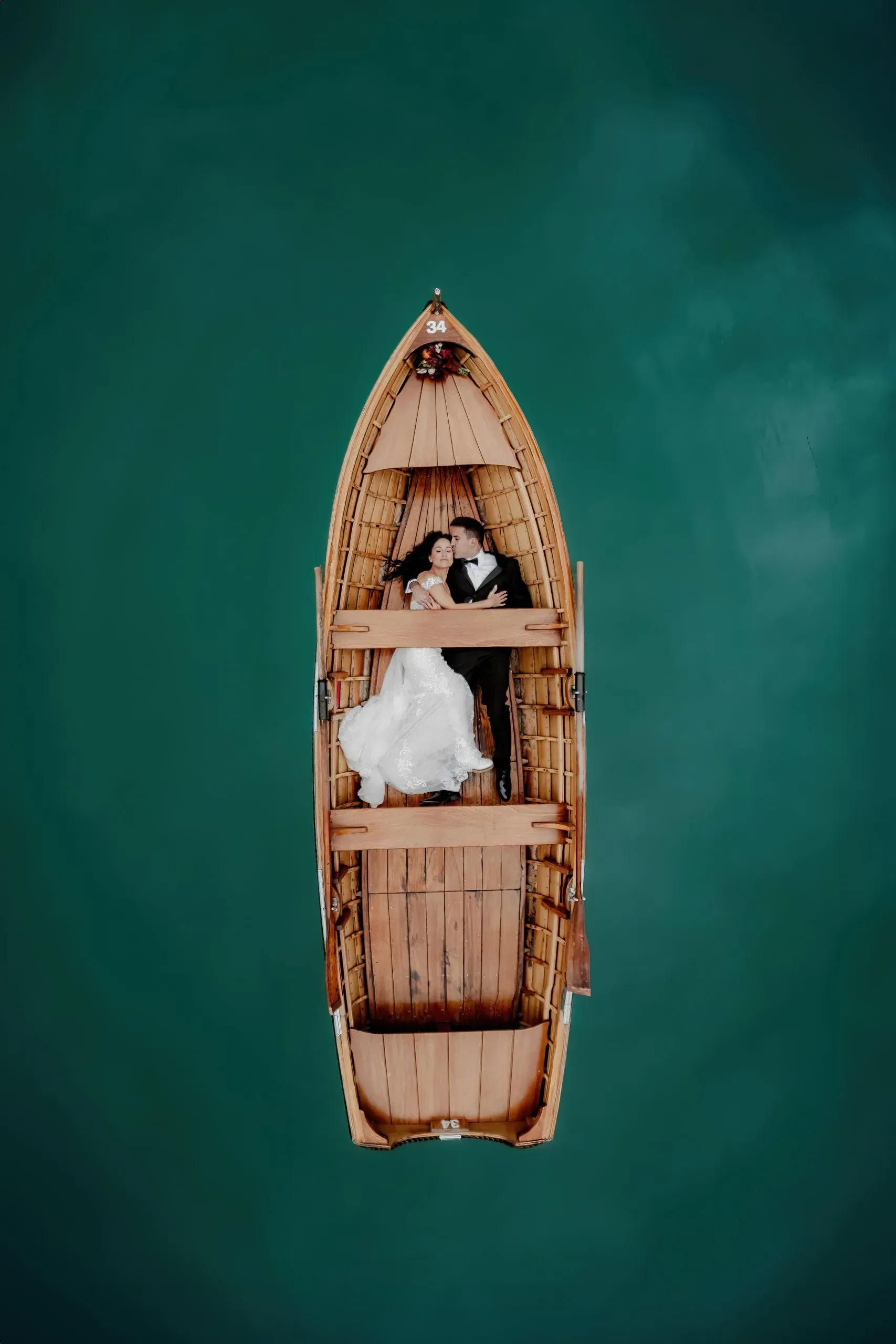 Romantisches Brautpaar liegt eng umschlungen in einem Holzboot auf türkisgrünem Wasser – aufgenommen aus der Vogelperspektive.