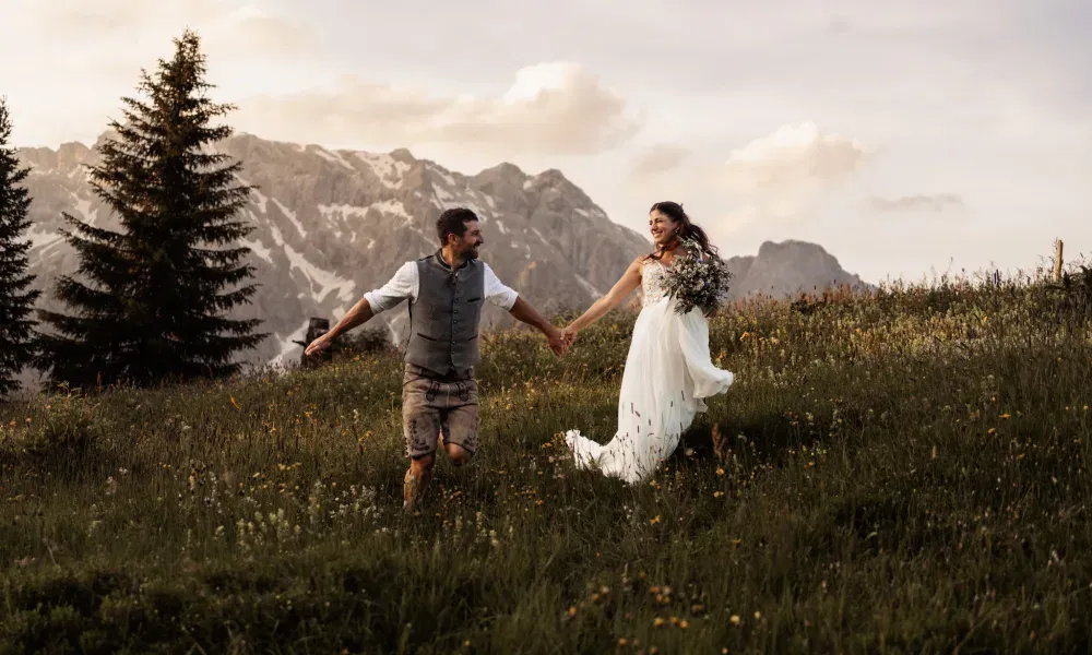 Fröhliches Brautpaar läuft Hand in Hand über eine blühende Almwiese vor imposanter Bergkulisse – festgehalten in warmem Abendlicht.