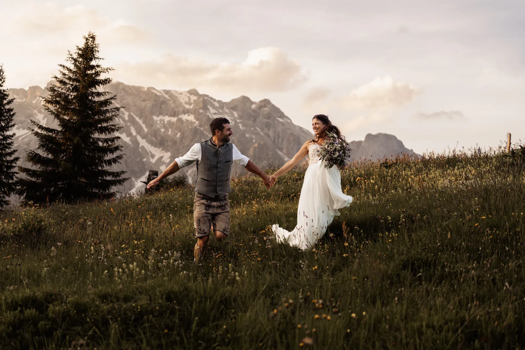 Fröhliches Brautpaar läuft Hand in Hand über eine blühende Almwiese vor imposanter Bergkulisse – festgehalten in warmem Abendlicht.