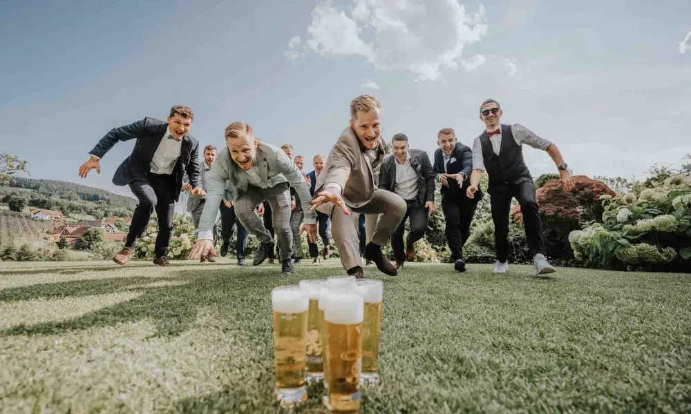 Ausgelassene Hochzeitsgesellschaft: Mehrere Männer in Anzügen rennen lachend über den Rasen auf eine Reihe Biergläser zu – ein humorvolles und energiegeladenes Gruppenfoto.