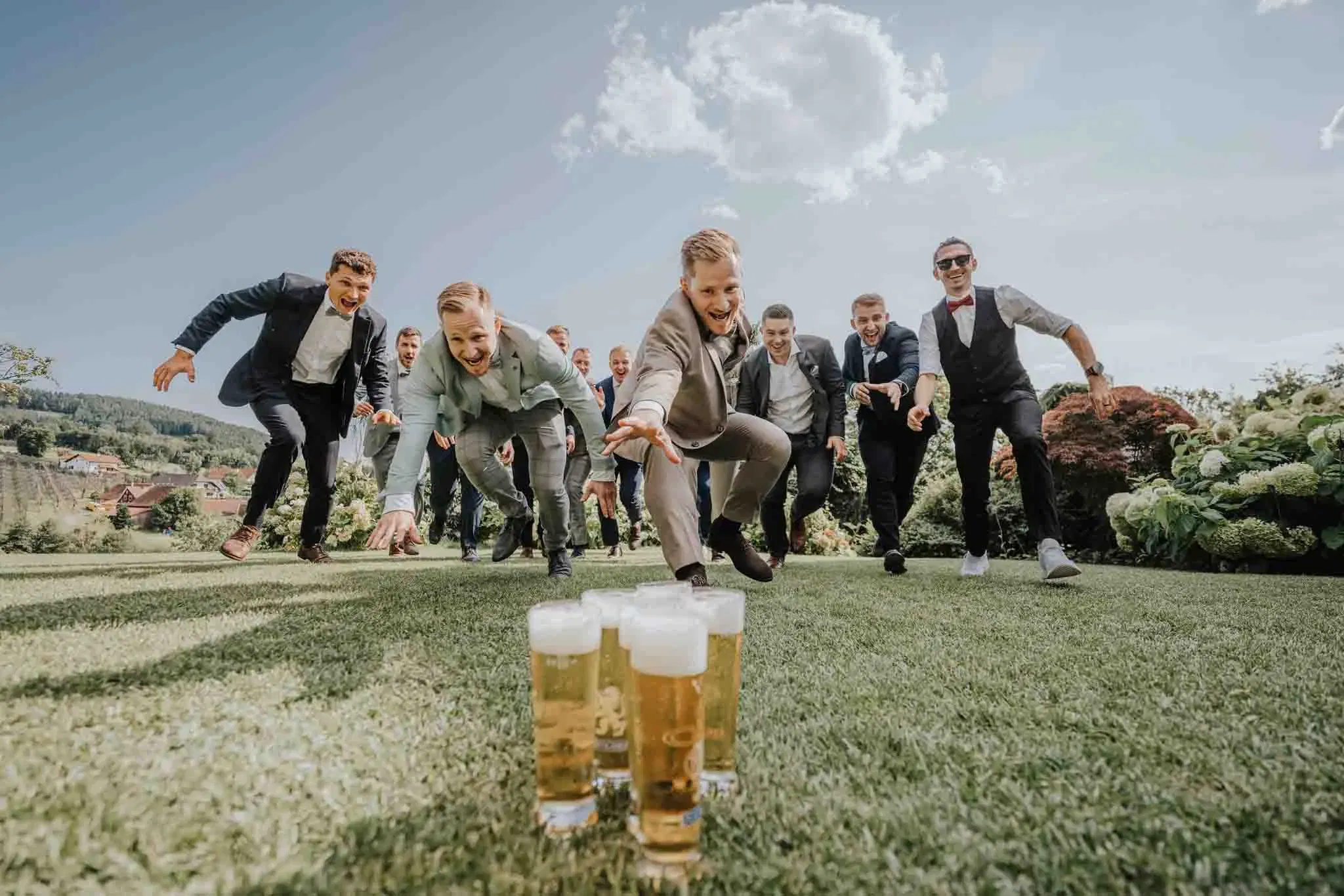 Ausgelassene Hochzeitsgesellschaft: Mehrere Männer in Anzügen rennen lachend über den Rasen auf eine Reihe Biergläser zu – ein humorvolles und energiegeladenes Gruppenfoto.