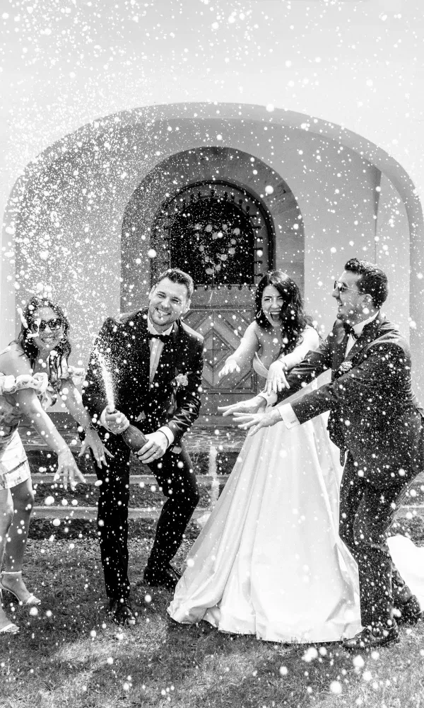 Ausgelassener Hochzeitsmoment: Das Brautpaar stößt gemeinsam mit Freunden an, während Champagner spritzend in die Luft steigt – festgehalten in lebendiger Schwarz-Weiß-Aufnahme.