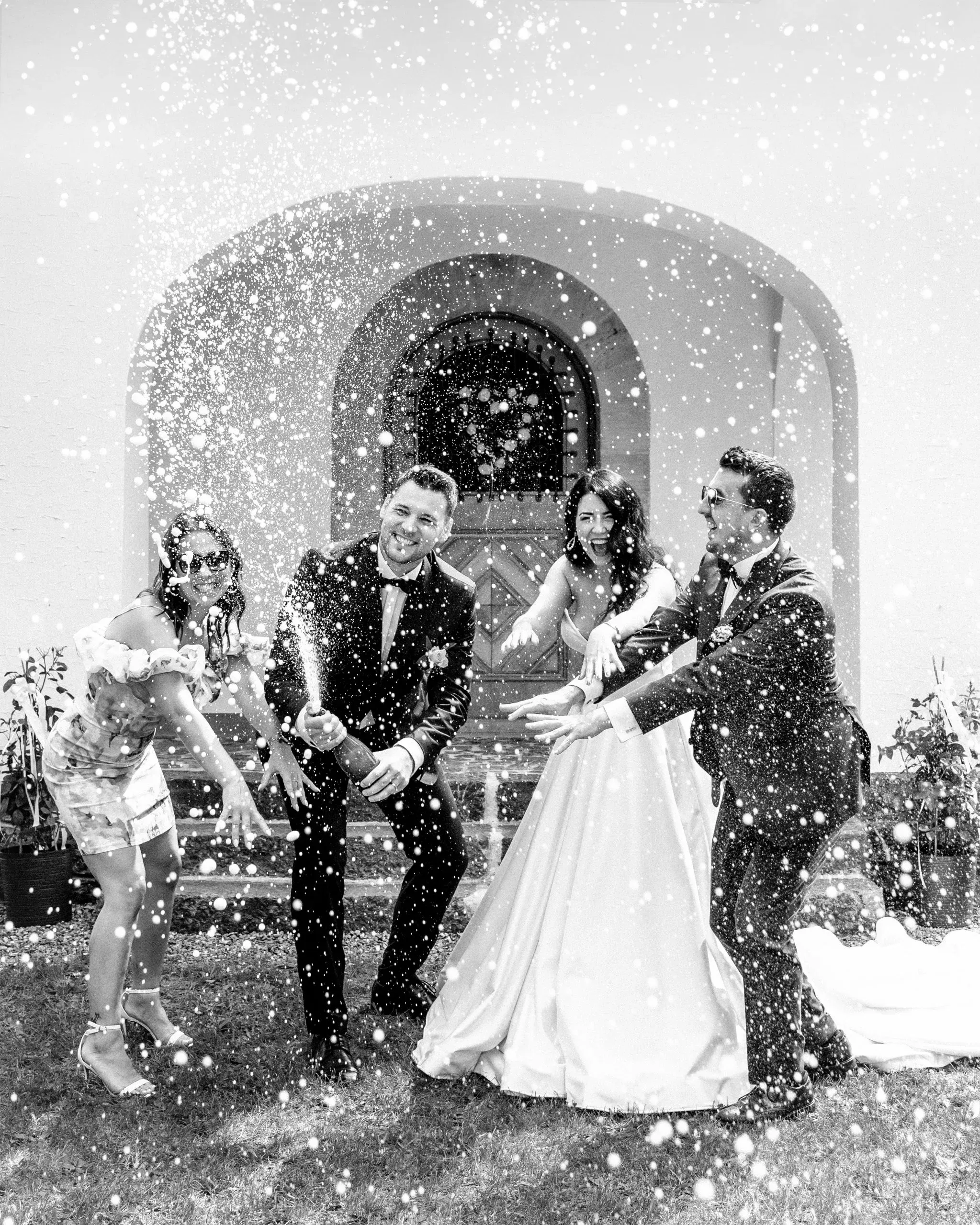Ausgelassener Hochzeitsmoment: Das Brautpaar stößt gemeinsam mit Freunden an, während Champagner spritzend in die Luft steigt – festgehalten in lebendiger Schwarz-Weiß-Aufnahme.