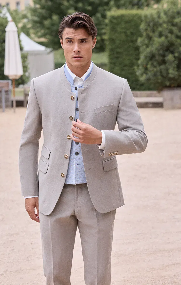 Bräutigam in beigem Hochzeitsanzug mit Stehkragen und messingfarbenen Knöpfen im modernen Trachtenstil von KUHN Maßkonfektion.