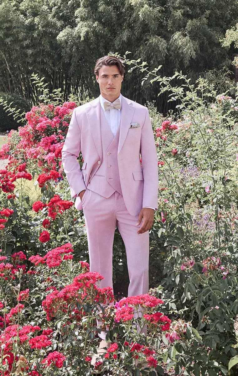 Bräutigam im roséfarbenen Dreiteiler mit Fliege, umgeben von blühenden Rosen – eleganter Hochzeitslook mit romantischem Flair.